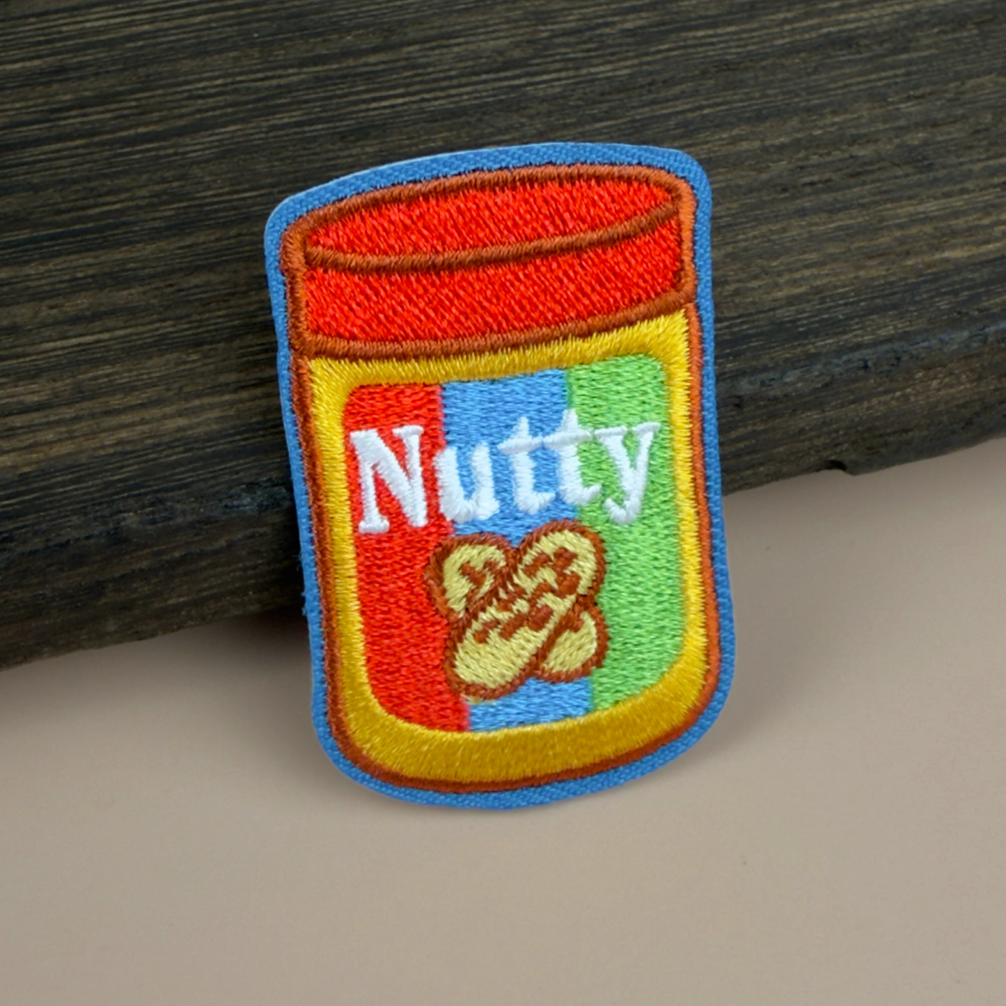 Embroidered patch of a peanut butter jar with 'Nutty' text on a wooden surface