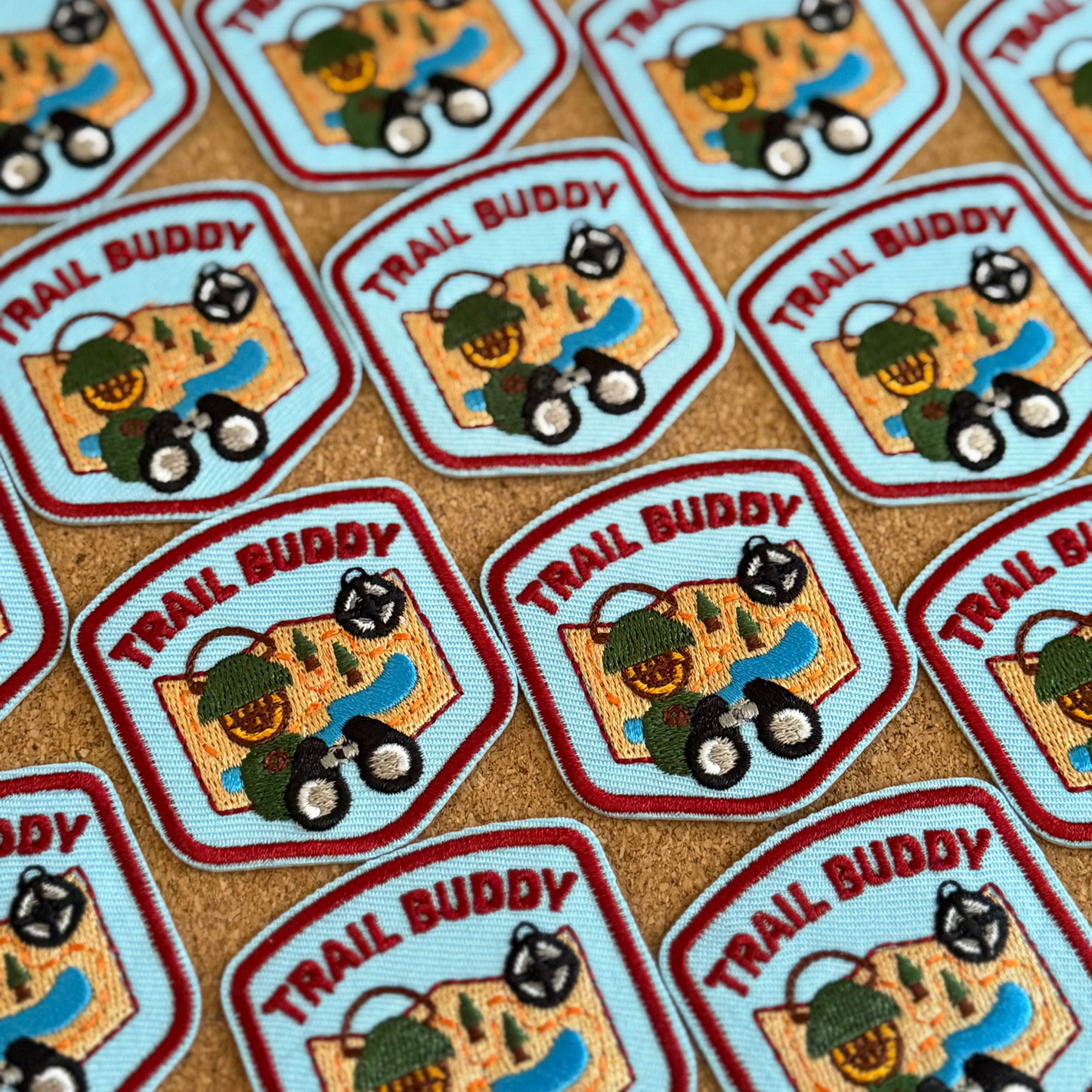 Multiple 'Trail Buddy' patches with a design of a camping lantern, binoculars, and a compass on a map on a brown background.