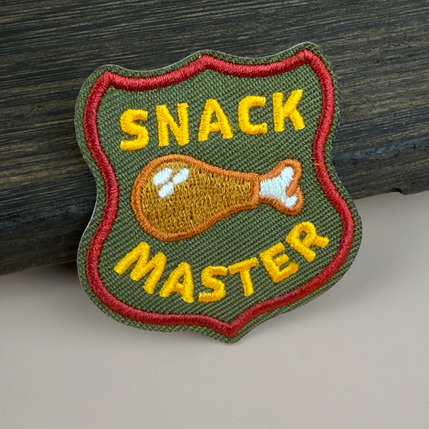 Embroidered patch with 'SNACK MASTER' text and a chicken drumstick design on a wooden background.