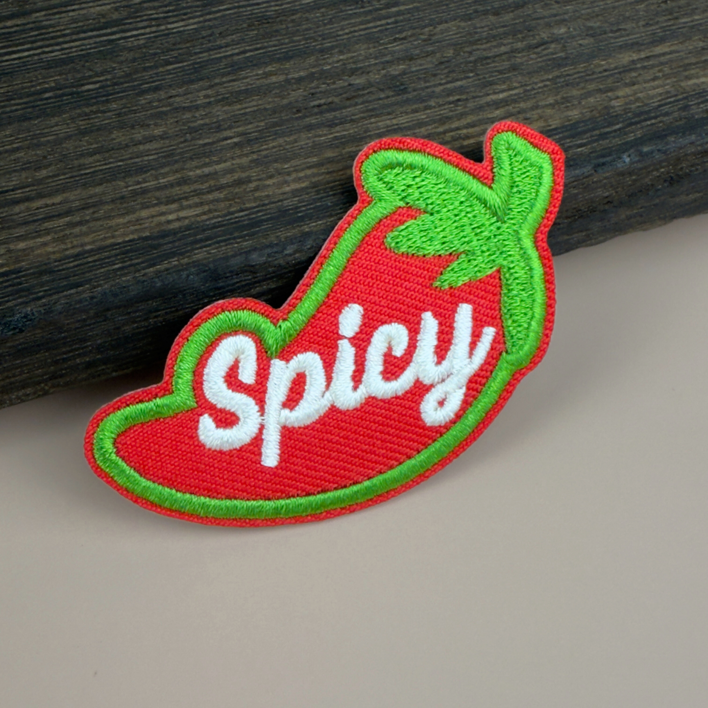 Embroidered patch with a red chili pepper design and 'Spicy' text on a wooden surface.