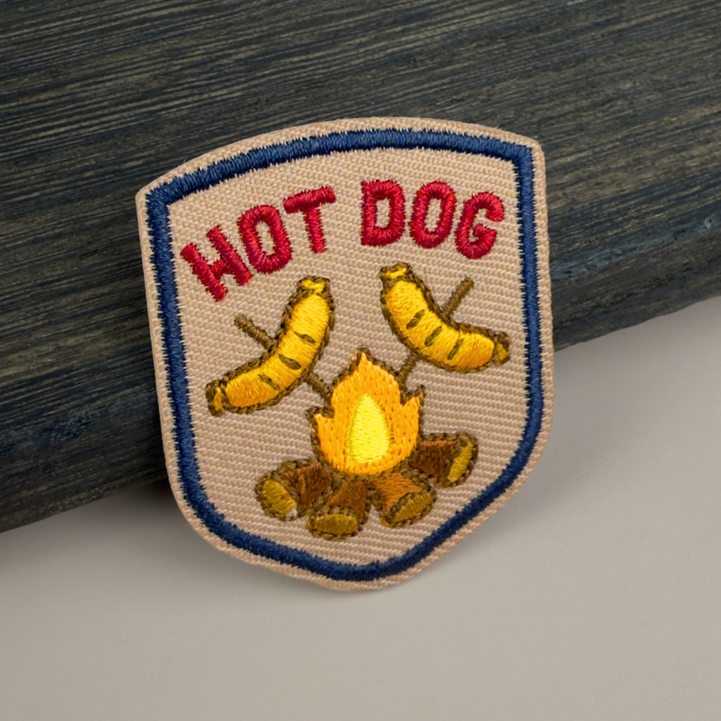 Embroidered patch with 'HOT DOG' text and two hot dogs on sticks cooking over a campfire design on a wooden surface