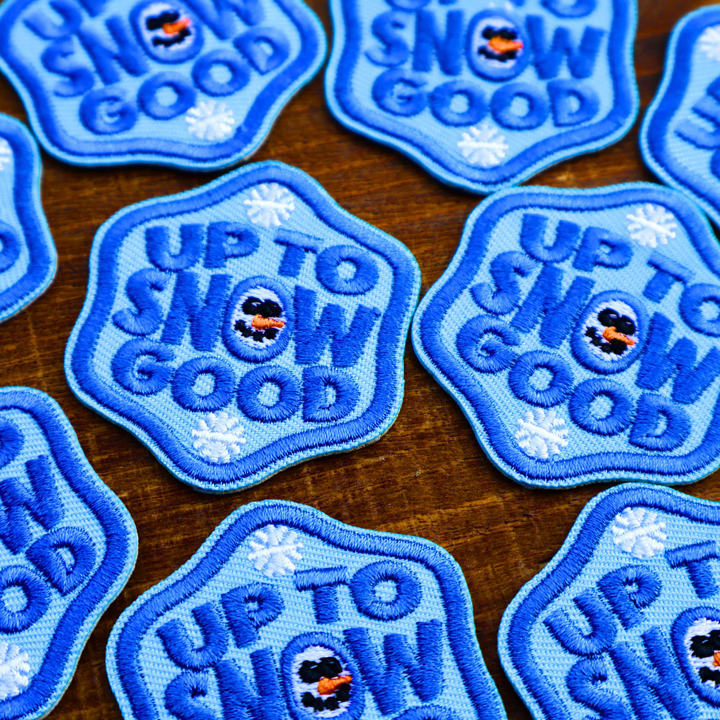 Blue iron-on patches with 'Up to Snow Good' text and snowman design on a wooden surface