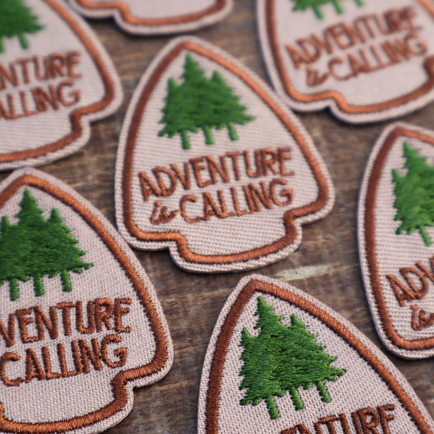 Embroidered iron-on patches with 'Adventure is Calling' text and green trees on a wooden surface.