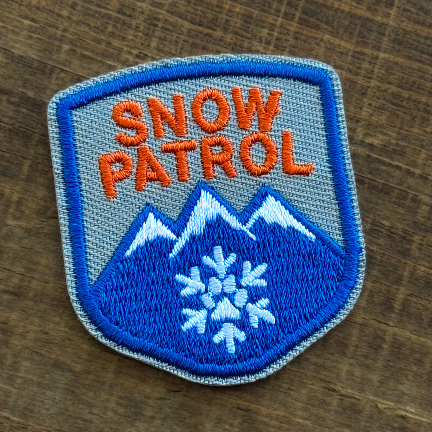 Iron-on embroidery patch for dogs with 'Snow Patrol' text and mountain design on a wooden surface