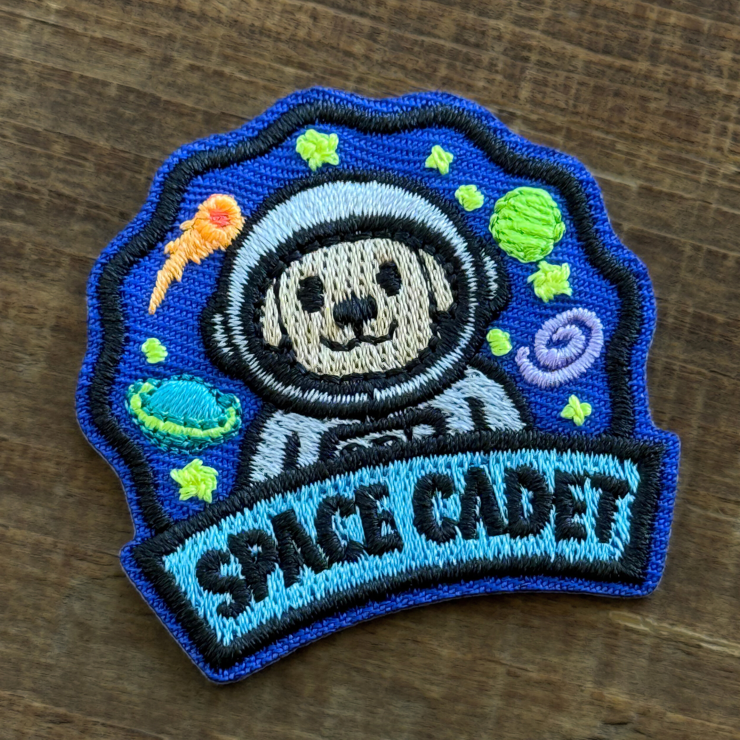 Embroidered iron-on patch of a dog in an astronaut suit with 'Space Cadet' text on a wooden surface