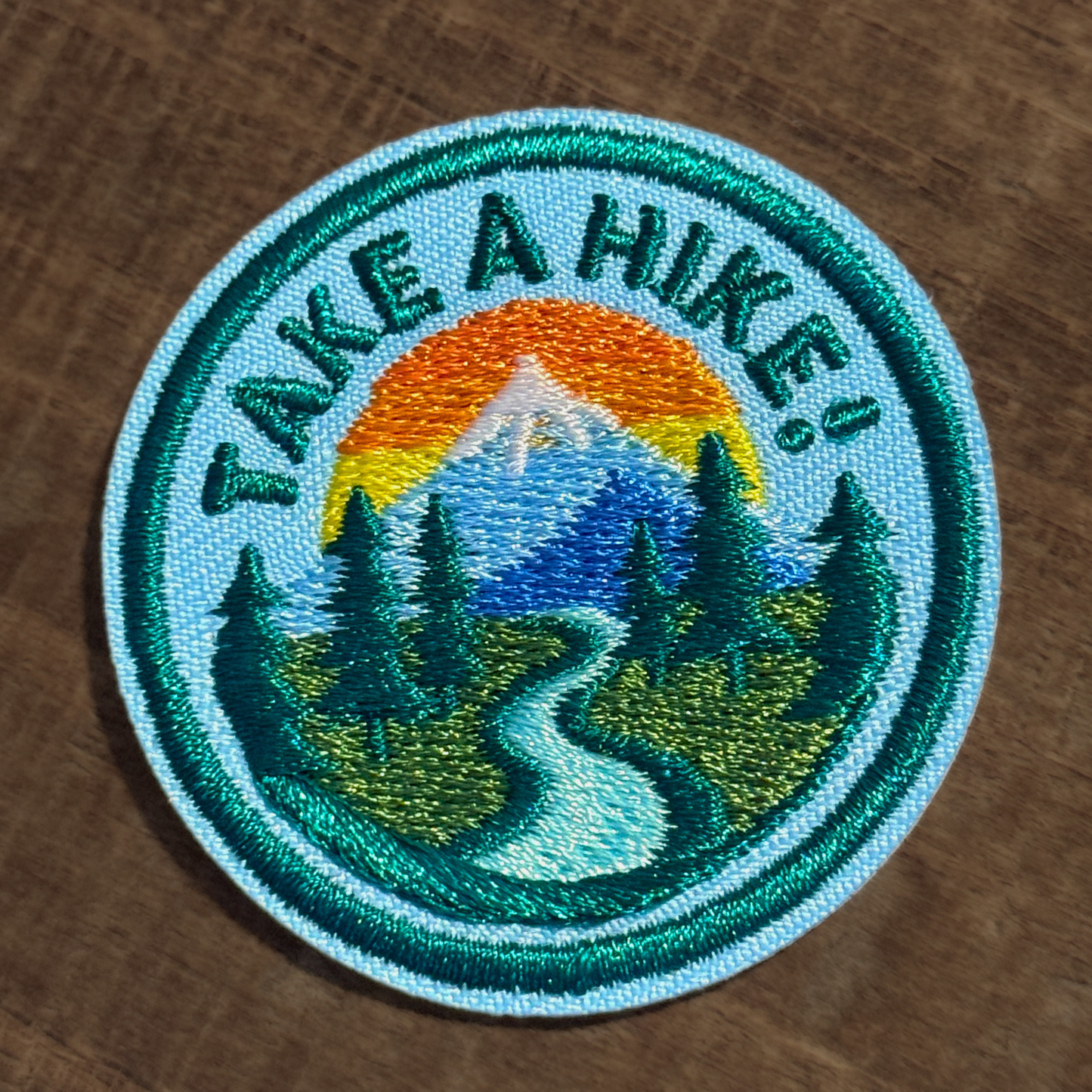 Embroidered iron-on patch for dogs with 'Take a Hike!' text and mountain landscape on a wooden surface