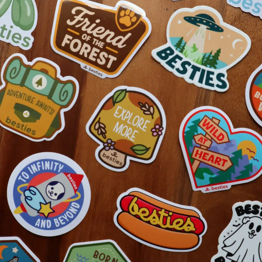 Flay lay of stickers arranged on a wooden surface.  Some stickers read as "Explore More" on a mushroom, "besties" written out in mustard on a got dog, and a Sci-Fi inspired design.